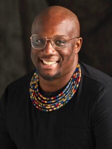 A headshot of Harold Steward, Executive Director at NEFA. He is facing the camera, smiling, wearing a black long sleeve, and adorning a colorful necklace.